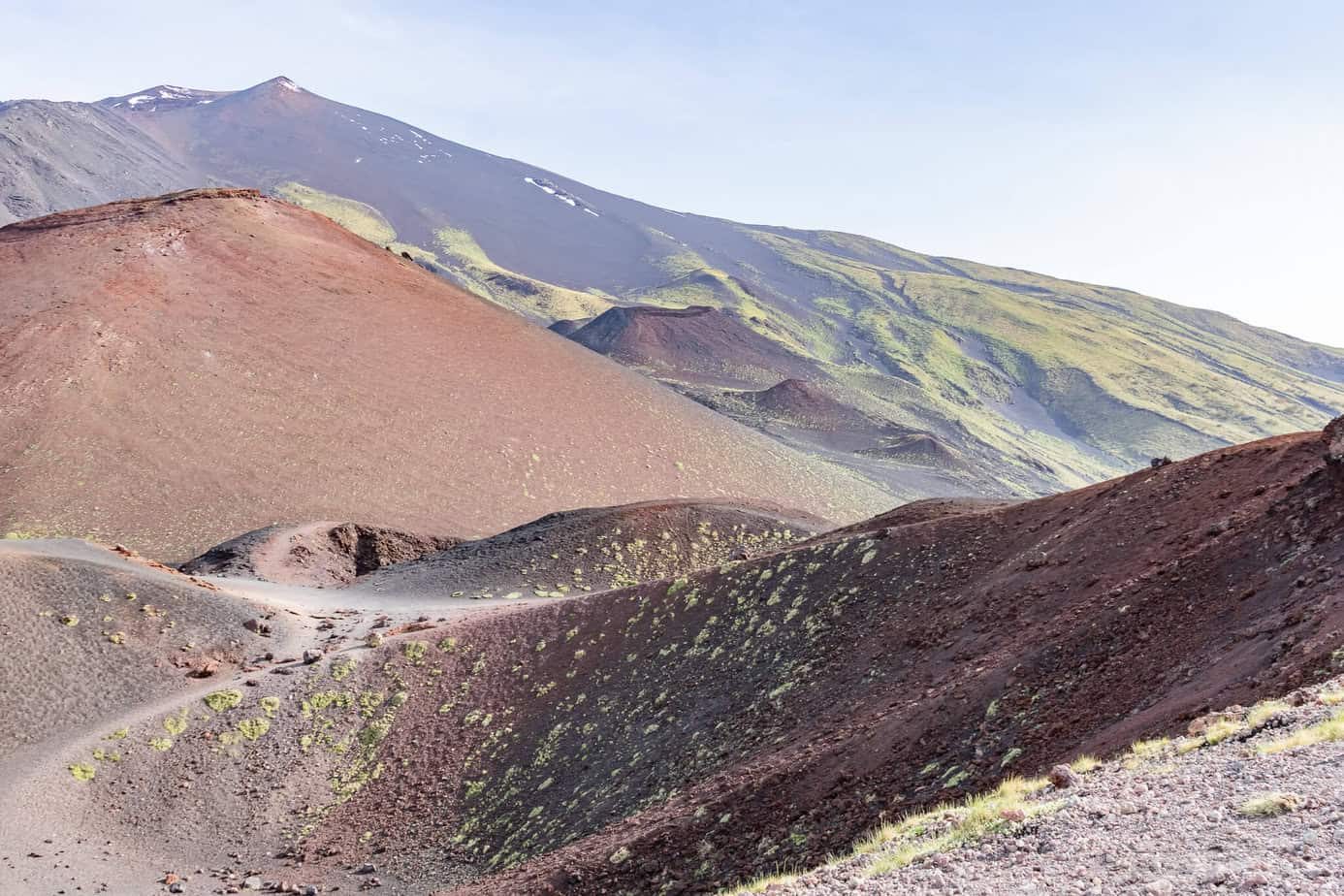 Eine Landschaftsansicht des Ätna mit rotbraunen Vulkanhängen im Vordergrund und einem größeren, grünen und felsigen Kegel im Hintergrund unter einem klaren Himmel.