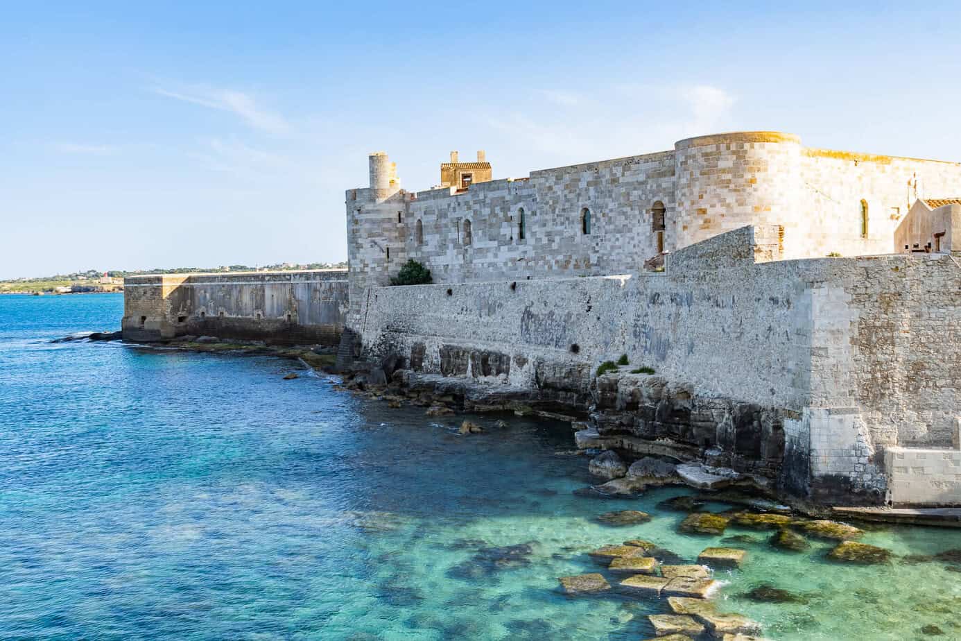 Die historische Festung in Ortigia steht am klaren, blaugrünen Meerwasser, mit felsiger Küste im Vordergrund und blauem Himmel darüber. Die Burgmauern reichen bis ans Wasser und schaffen so eine malerische Küstenkulisse.
