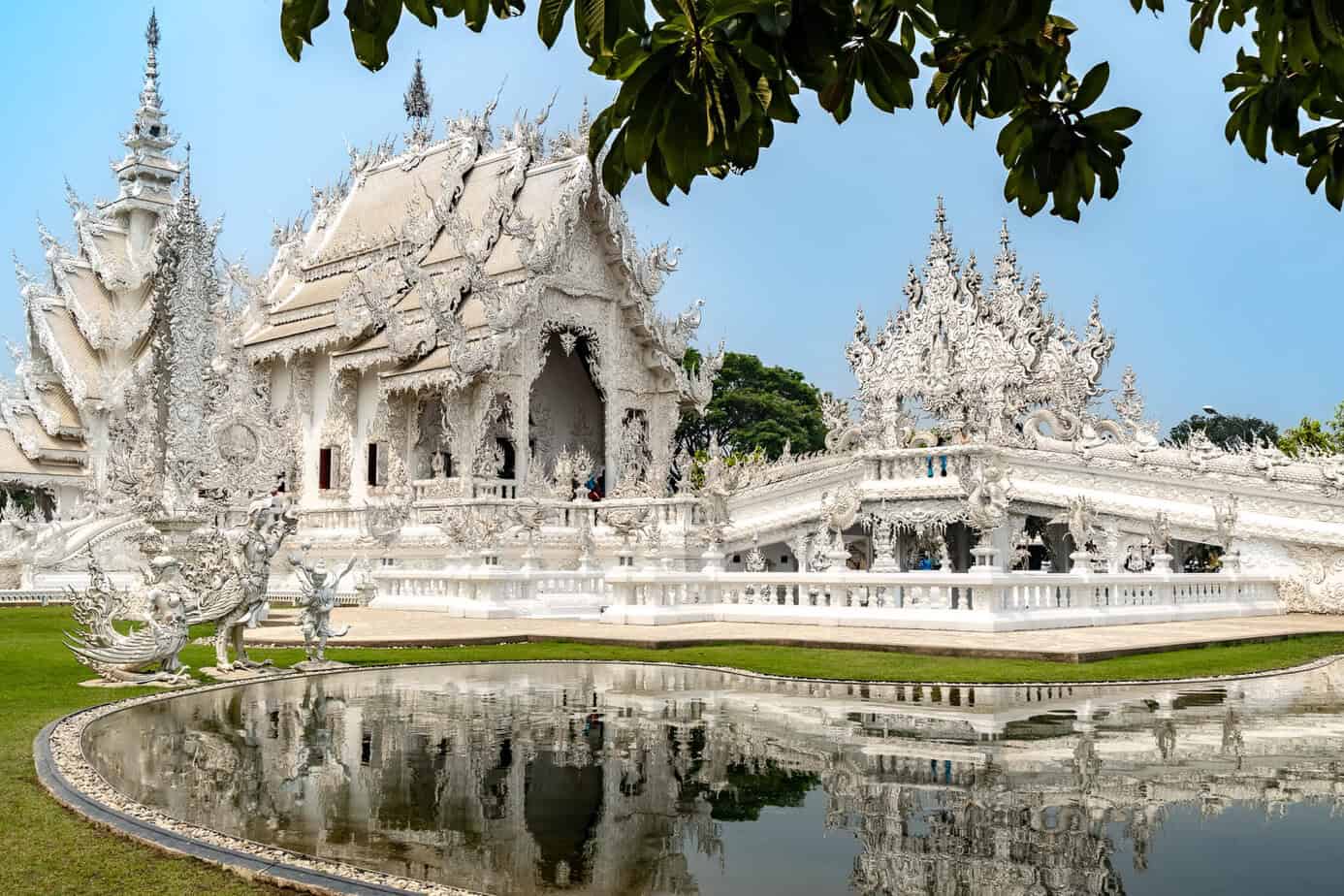 Der kunstvoll verzierte White Temple mit Spiegelmosaiken, der sich im stillen Teich spiegelt, umgeben von Grün und überhängenden Ästen unter einem klaren blauen Himmel.