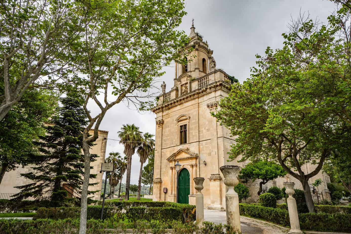 Historische Steinkirche im Giardino Ibleo mit markantem Glockenturm und grünen Türen, umgeben von üppigen Bäumen und gepflegten Büschen. Perfekt zum Schlendern!