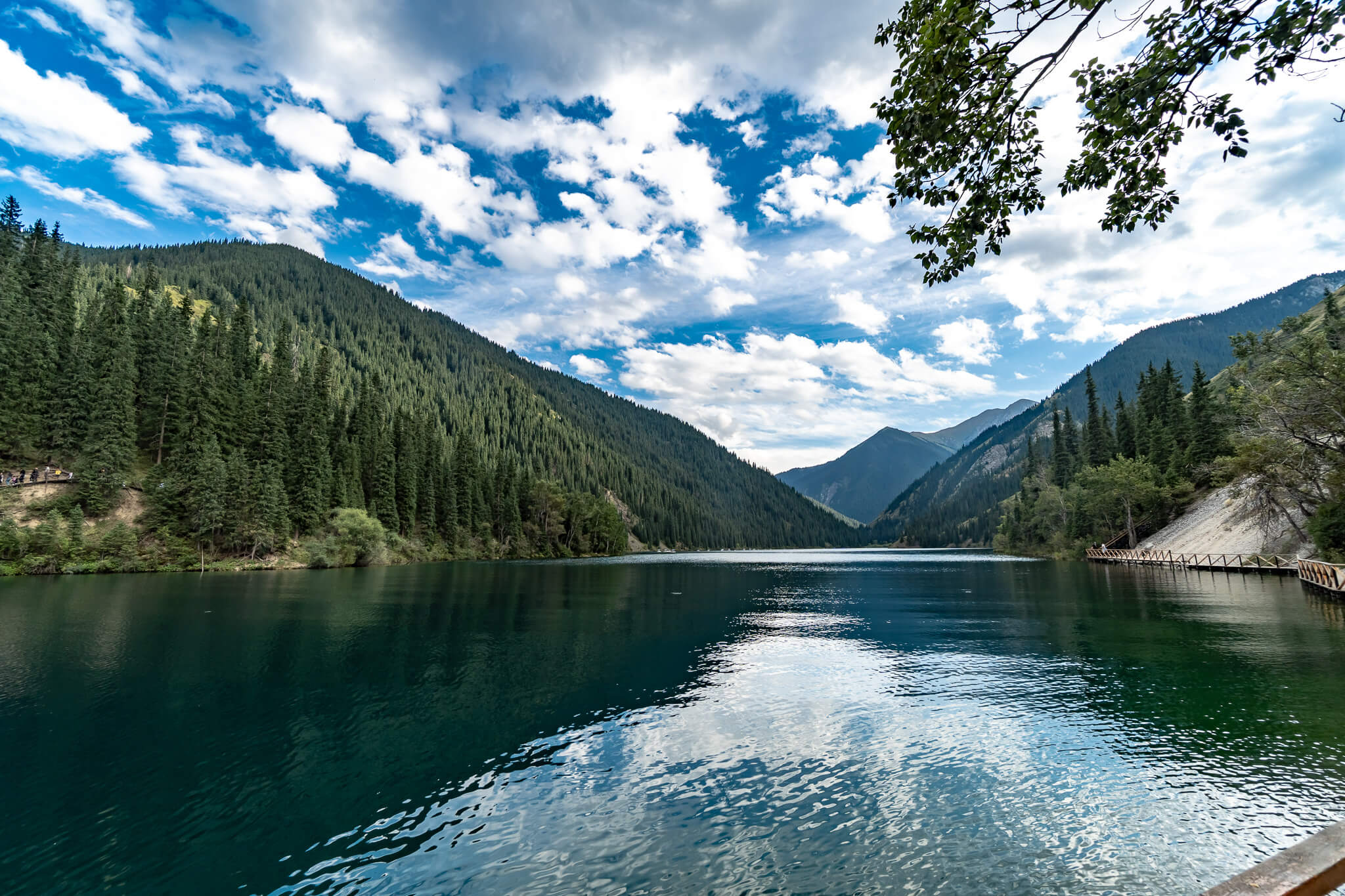 Almaty Sehenswürdigkeiten: Die schönsten Highlights & meine 8 Tipps 18 Dunkelgrüner See mit spiegelnden Wolken, umgeben von Nadelwald und Bergen, rechts ein Holzsteg am Ufer