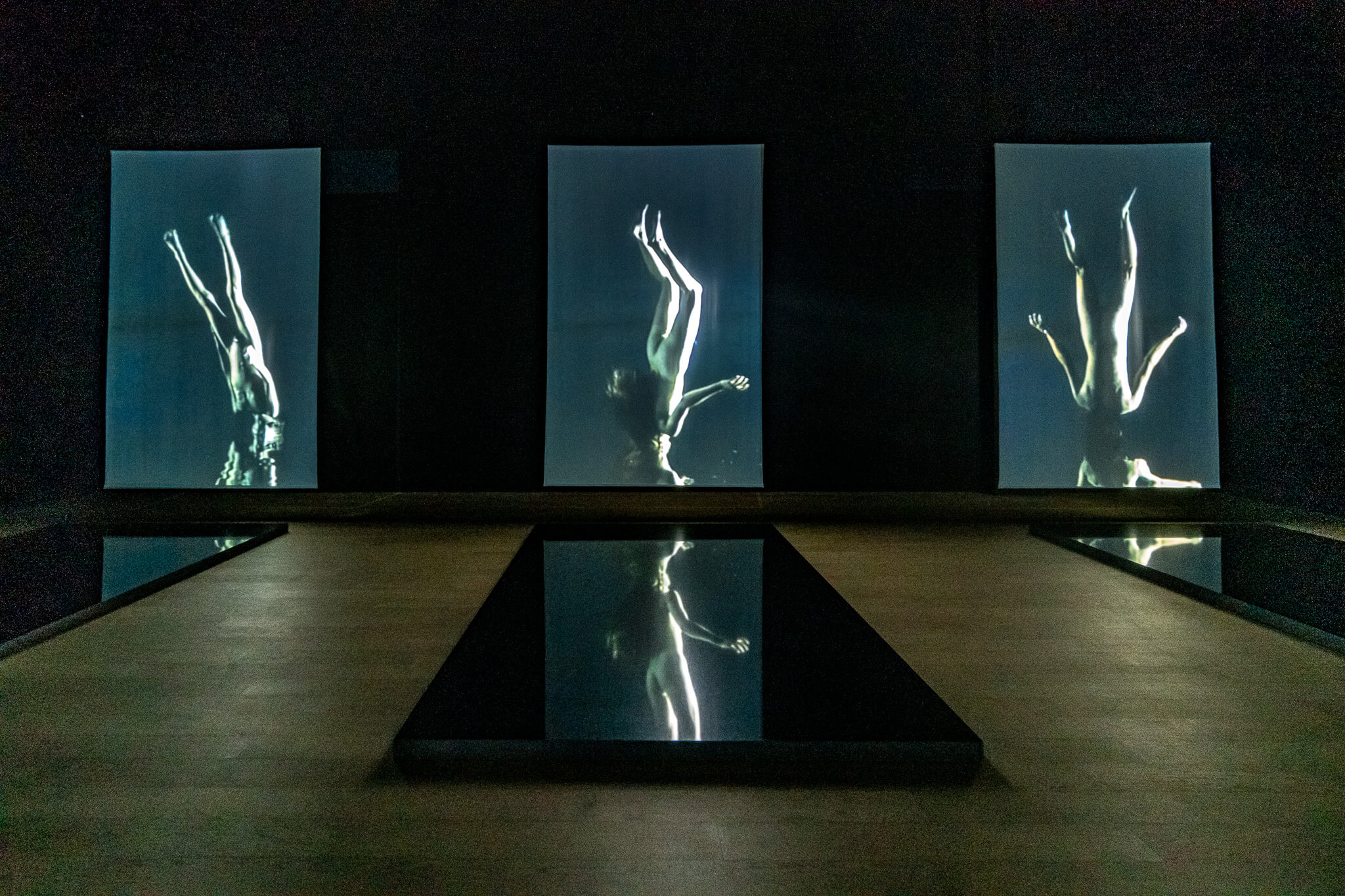 Almaty Sehenswürdigkeiten: Die schönsten Highlights & meine 8 Tipps 11 Dunkler Ausstellungsraum mit drei hochformatigen Videoleinwänden mit kopfüber schwebenden Figuren im Wasser, davor spiegelnde Becken