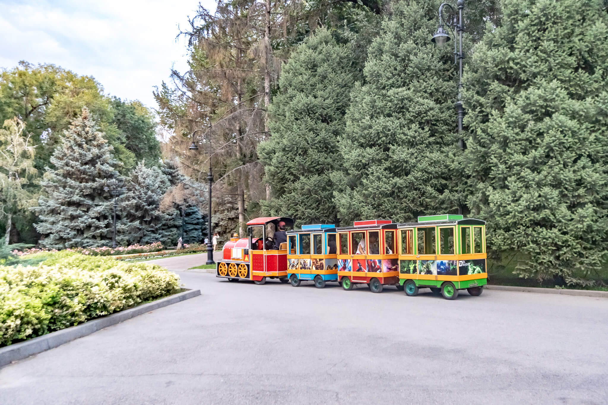 Almaty Sehenswürdigkeiten: Die schönsten Highlights & meine 8 Tipps 6 Bunte Touristenbahn auf einem Weg im Park der 28 Panfilowzy in Almaty, umgeben von dichten Nadelbäumen und Hecken